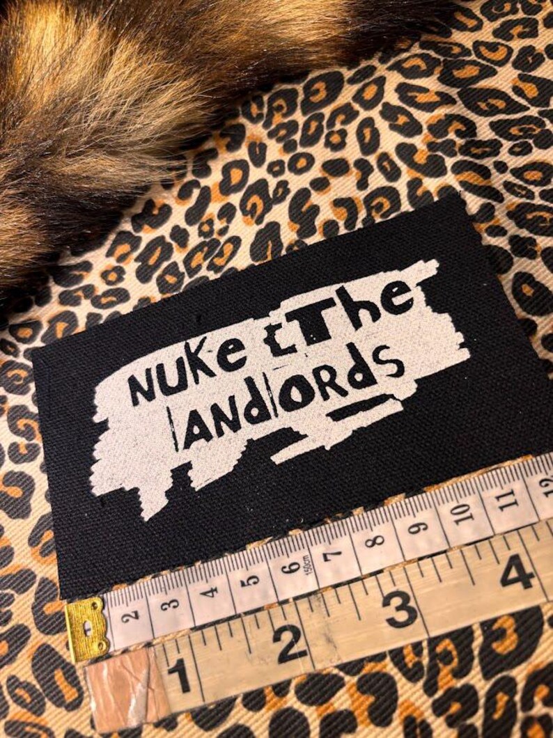 Peut inclure: &Eacute;cusson rectangulaire noir avec le texte "Nuke the Landlords" en blanc. Un m&egrave;tre ruban indique que l'&eacute;cusson mesure environ 10 cm de large. Le fond pr&eacute;sente un motif l&eacute;opard.
