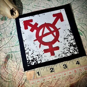 May include: A square fabric patch featuring a red anarchy symbol combined with gender symbols on a white background with black distressed edges. A yellow measuring tape is placed along the bottom edge, showing a width of approximately 4 inches.