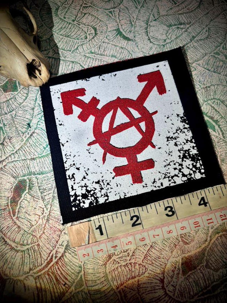 May include: A square fabric patch featuring a red anarchy symbol combined with gender symbols on a white background with black distressed edges. A yellow measuring tape is placed along the bottom edge, showing a width of approximately 4 inches.