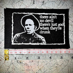 Peut inclure: Patch brodé noir et blanc avec un portrait d'un homme portant un chapeau et le texte "there ain't no devil there's just god when they're drunk".