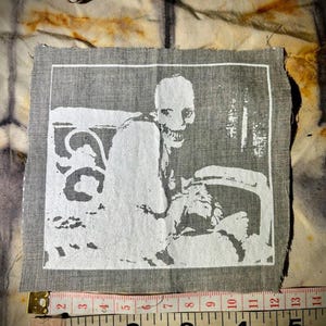 Puede incluir: Un parche de tela gris con una ilustraci&oacute;n blanca de un esqueleto sentado en una silla. La imagen est&aacute; enmarcada en blanco. El parche mide aproximadamente 10 cm de ancho, como muestra la cinta m&eacute;trica. El fondo es una tela texturizada y estampada.