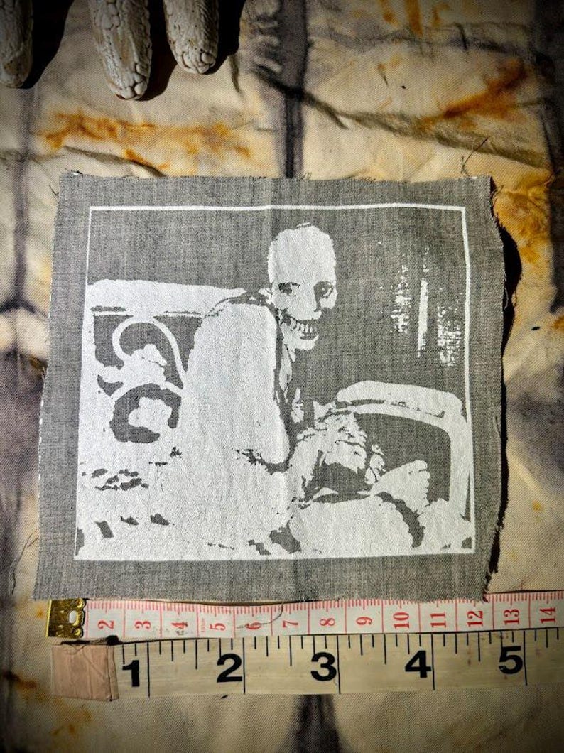 Puede incluir: Un parche de tela gris con una ilustraci&oacute;n blanca de un esqueleto sentado en una silla. La imagen est&aacute; enmarcada en blanco. El parche mide aproximadamente 10 cm de ancho, como muestra la cinta m&eacute;trica. El fondo es una tela texturizada y estampada.