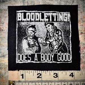 May include: Black and white embroidered patch with the text "BLOODLETTING! DOES A BODY GOOD" and a graphic of two men, one holding a knife, the other with a bandage on their arm.