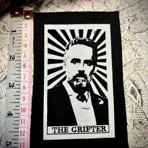 Può includere: Toppa termoadesiva in bianco e nero con un'immagine stilizzata di un viso maschile con barba e baffi. L'immagine è circondata da un design a raggiera. Il testo "THE GRIFTER" è stampato sotto l'immagine.