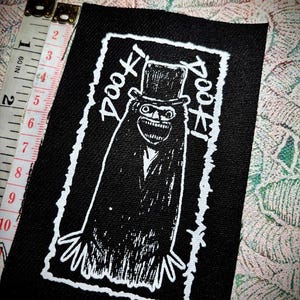 May include: Black rectangular patch with a white illustration of a creature wearing a top hat and the text "DOOF!" in a stylized font. The patch measures approximately 3 inches in height and 2 inches in width.