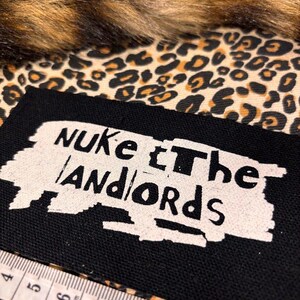 Peut inclure: Un patch noir avec le texte "NUKE the LANDLORDS" en blanc, sur un fond imprim&eacute; l&eacute;opard et une bordure en fausse fourrure marron. Un m&egrave;tre ruban est au premier plan.