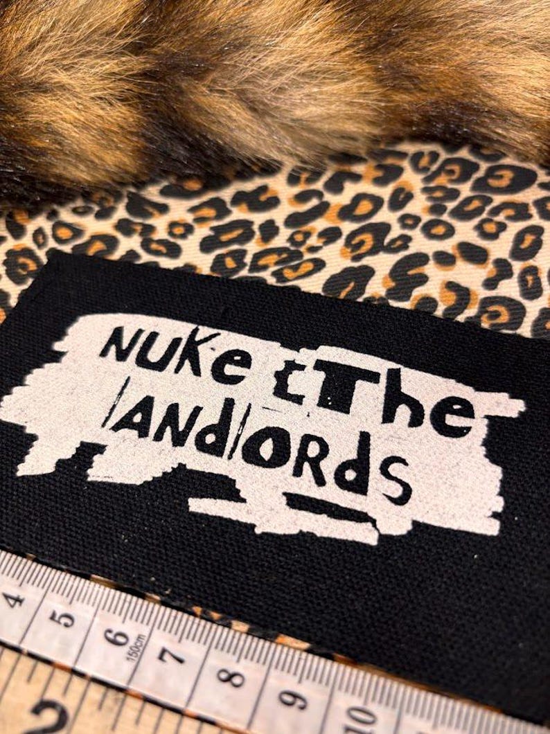 Peut inclure: Un patch noir avec le texte "NUKE the LANDLORDS" en blanc, sur un fond imprim&eacute; l&eacute;opard et une bordure en fausse fourrure marron. Un m&egrave;tre ruban est au premier plan.