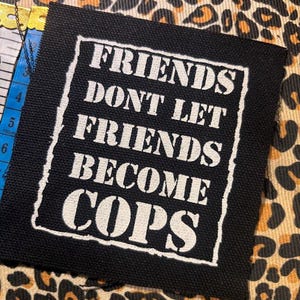 May include: A square black fabric patch featuring white screen-printed text that reads FRIENDS DONT LET FRIENDS BECOME COPS, enclosed within a white rectangular border. A yellow measuring tape is visible, showing a length of approximately 6 inches.