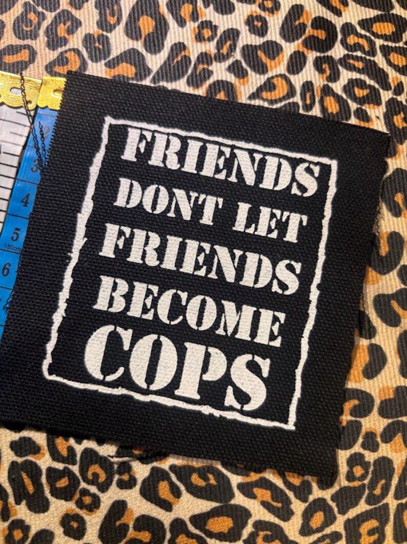 May include: A square black fabric patch featuring white screen-printed text that reads FRIENDS DONT LET FRIENDS BECOME COPS, enclosed within a white rectangular border. A yellow measuring tape is visible, showing a length of approximately 6 inches.