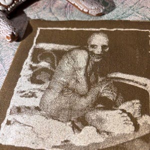 Puede incluir: Un parche de tela marr&oacute;n con una imagen en tonos sepia de una figura sentada con un rostro esquel&eacute;tico. La imagen est&aacute; enmarcada por un borde blanco. Un objeto blanco texturizado es parcialmente visible en el fondo.