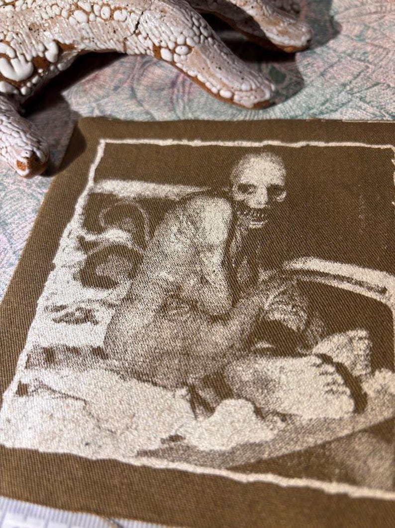 Puede incluir: Un parche de tela marr&oacute;n con una imagen en tonos sepia de una figura sentada con un rostro esquel&eacute;tico. La imagen est&aacute; enmarcada por un borde blanco. Un objeto blanco texturizado es parcialmente visible en el fondo.