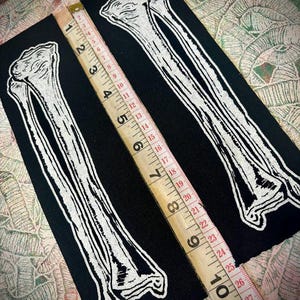 Op de afbeelding: Twee zwarte patches met witte botillustraties. Een houten meetlint, gemarkeerd in inches, ligt over de patches. De patches zijn ongeveer 25 cm lang.