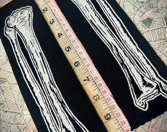 Puede incluir: Dos parches negros con ilustraciones de huesos blancos. Una cinta métrica de madera, marcada en pulgadas, se coloca sobre los parches. Los parches miden aproximadamente 25 cm de largo.