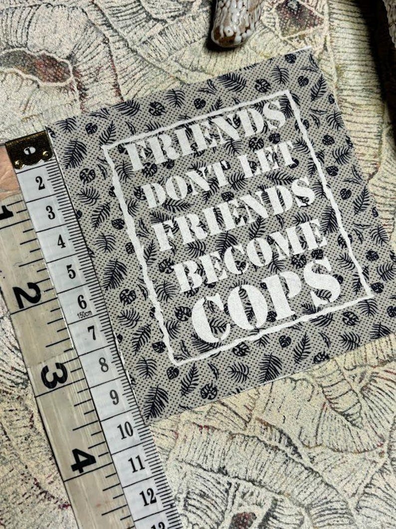 May include: A square fabric patch with a beige background and a black botanical leaf pattern. The text "FRIENDS DON'T LET FRIENDS BECOME COPS" is printed in white in the center. A measuring tape shows the patch is approximately 4 inches wide.