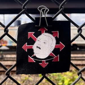 Puede incluir: Un parche de tela negro cuadrado que presenta una ilustración de líneas blancas de la cabeza de un personaje con expresión de sorpresa, centrada sobre un símbolo de estrella roja de ocho puntas con flechas hacia afuera, sujeto con un clip.