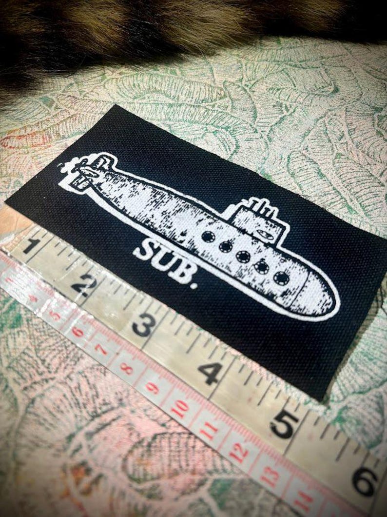 May include: A rectangular black fabric patch featuring a white screen-printed illustration of a submarine with the text SUB. positioned below it. A measuring tape is placed along the bottom edge, showing a length of approximately 11.5 centimetres.