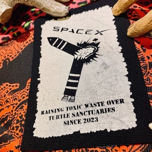May include: A rectangular black and white fabric patch featuring the text SPACEX at the top, a graphic of a rocket emitting a plume of smoke, and the text RAINING TOXIC WASTE OVER TURTLE SANCTUARIES SINCE 2023 printed in black on a white background.