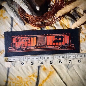May include: A rectangular black patch featuring an orange illustration of a Burlington Northern train car. The patch is approximately 5 inches long. The text "BURLINGTON NORTHERN" and "BN 355145" are visible. A measuring tape is placed below the patch.