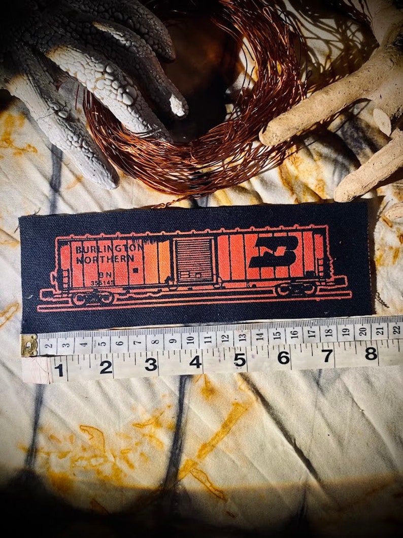 May include: A rectangular black patch featuring an orange illustration of a Burlington Northern train car. The patch is approximately 5 inches long. The text "BURLINGTON NORTHERN" and "BN 355145" are visible. A measuring tape is placed below the patch.