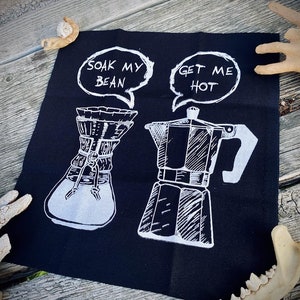 May include: Black fabric patch with white text and illustrations. The text reads "Soak My Bean" and "Get Me Hot". The illustrations are of a coffee maker and a pour-over coffee brewer.