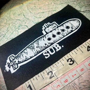 May include: A rectangular black fabric patch featuring a white screen-printed graphic of a submarine with the text SUB. positioned below it. A white measuring tape is placed along the bottom edge, showing a length of approximately 8.9 centimetres.