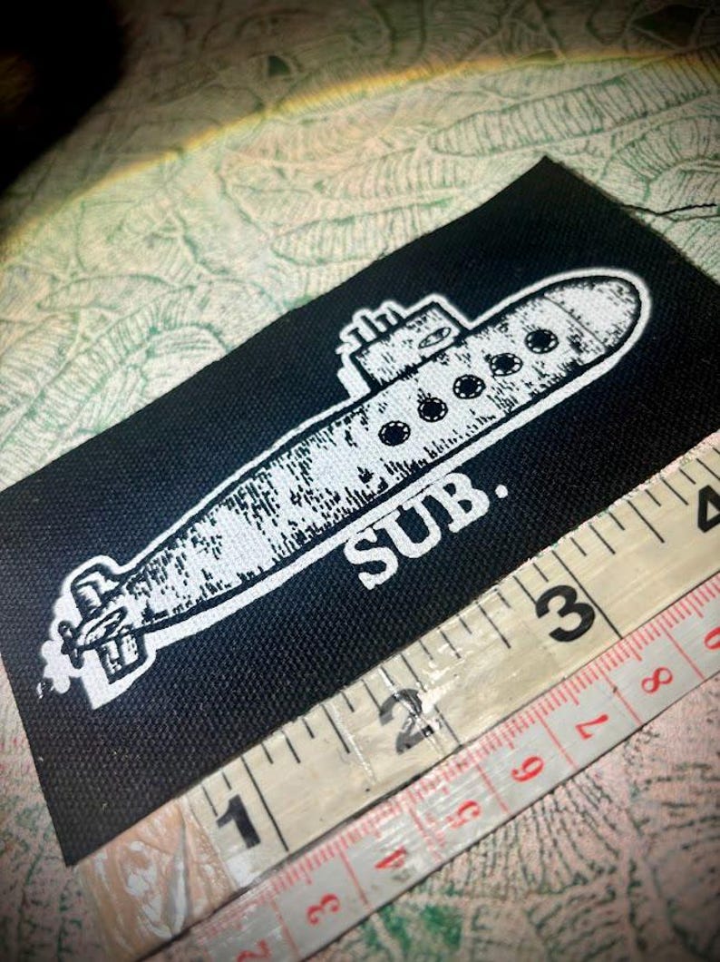 May include: A rectangular black fabric patch featuring a white screen-printed graphic of a submarine with the text SUB. positioned below it. A white measuring tape is placed along the bottom edge, showing a length of approximately 8.9 centimetres.
