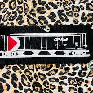 Op de afbeelding: Zwart-witte patch van een CP Rail goederenwagon met een rode driehoek aan de zijkant. De patch bevindt zich op een achtergrond van luipaardprintstof.