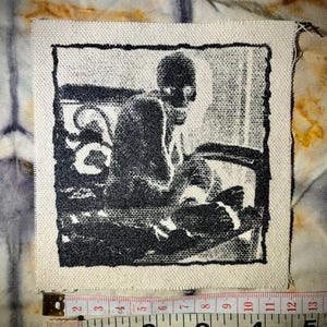 Puede incluir: Parche de tela cuadrado con una ilustraci&oacute;n en blanco y negro de una persona sentada en una mesa. El parche mide aproximadamente 10 cm de ancho. Los bordes no est&aacute;n acabados, sobre un fondo te&ntilde;ido anudado.