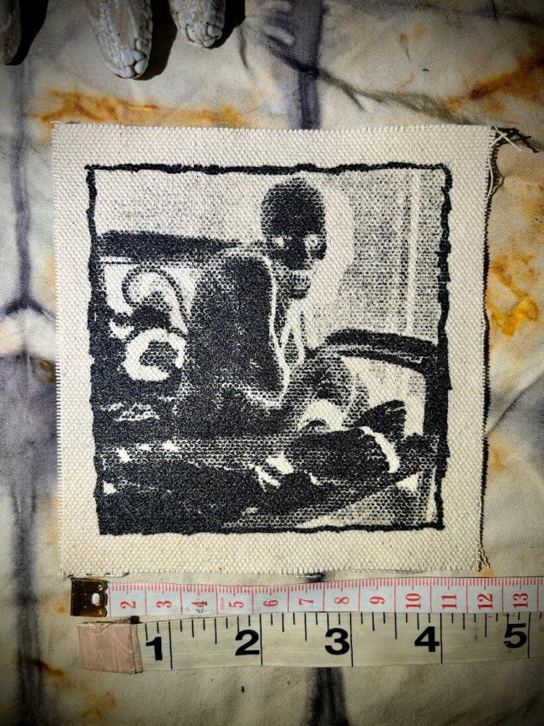 Puede incluir: Parche de tela cuadrado con una ilustraci&oacute;n en blanco y negro de una persona sentada en una mesa. El parche mide aproximadamente 10 cm de ancho. Los bordes no est&aacute;n acabados, sobre un fondo te&ntilde;ido anudado.