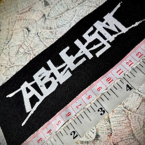 Peut inclure: Un patch en tissu noir avec le mot "ABUSEMENT" écrit en lettres blanches inclinées. Le patch est mesuré avec un mètre ruban blanc, montrant 7,6 cm.