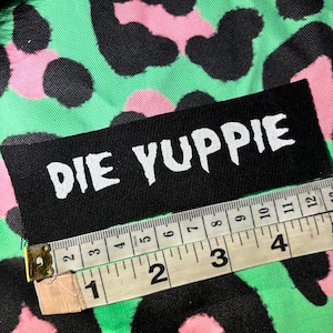 May include: Black fabric patch with white text that reads "DIE YUPPIE". The patch is lying on a green and pink leopard print fabric. A measuring tape is partially visible in the bottom right corner of the image.