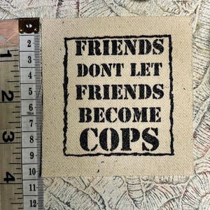 May include: A square beige fabric patch featuring the black text "FRIENDS DONT LET FRIENDS BECOME COPS" inside a black rectangular border. A measuring tape is positioned along the left side, showing the patch is approximately 4 inches tall.