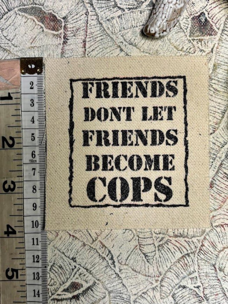 May include: A square beige fabric patch featuring the black text "FRIENDS DONT LET FRIENDS BECOME COPS" inside a black rectangular border. A measuring tape is positioned along the left side, showing the patch is approximately 4 inches tall.