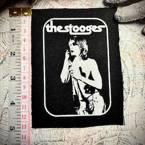 May include: Black and white patch featuring the band name "the stooges" in a white rounded rectangle. The patch has a black background with a white image of a person holding a microphone. The patch measures approximately 4.5 inches in height and 3.5 inches in width.
