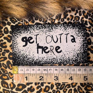 Peut inclure: Un écusson rectangulaire avec les mots "get OUTTA here" en noir sur fond blanc avec une bordure mouchetée. L'écusson est sur un tissu à imprimé léopard. Une règle indique que l'écusson mesure environ 10 cm de large.