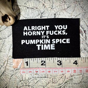 May include: A black rectangular patch with white text that reads "ALRIGHT YOU HORNY FUCKS, IT'S PUMPKIN SPICE TIME." A ruler is placed below the patch, showing measurements in inches.