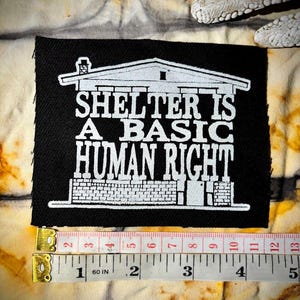 Op de afbeelding: Zwarte rechthoekige patch met witte tekst: "SHELTER IS A BASIC HUMAN RIGHT." De patch heeft een huisafbeelding. De patch is ongeveer 7,6 cm breed, zoals te zien is op de meetlint.