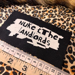 Peut inclure: &Eacute;cusson rectangulaire noir avec le texte "Nuke the Landlords" en blanc. Une r&egrave;gle en bois avec des mesures en centim&egrave;tres est plac&eacute;e &agrave; c&ocirc;t&eacute; de l'&eacute;cusson. Le fond pr&eacute;sente un motif l&eacute;opard.