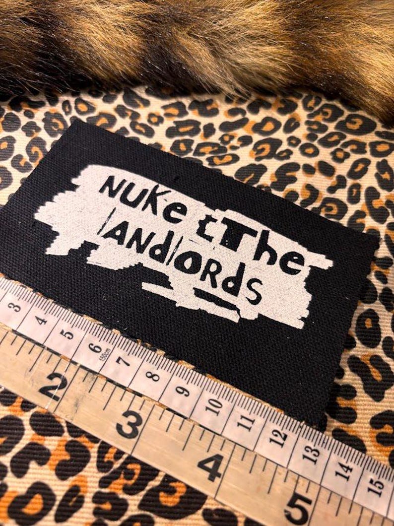 Peut inclure: &Eacute;cusson rectangulaire noir avec le texte "Nuke the Landlords" en blanc. Une r&egrave;gle en bois avec des mesures en centim&egrave;tres est plac&eacute;e &agrave; c&ocirc;t&eacute; de l'&eacute;cusson. Le fond pr&eacute;sente un motif l&eacute;opard.