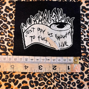 May include: Black square patch featuring a white graphic of a toilet roll and text. The text reads "JUST PAY US ENOUGH TO FUCK LIVE". The patch measures approximately 7.6 cm wide.
