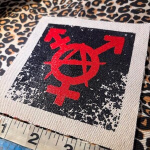 May include: A square fabric patch featuring a red anarchy symbol combined with gender symbols printed on a black distressed background. A white measuring tape is positioned along the bottom edge, showing the patch width is approximately 4 inches.