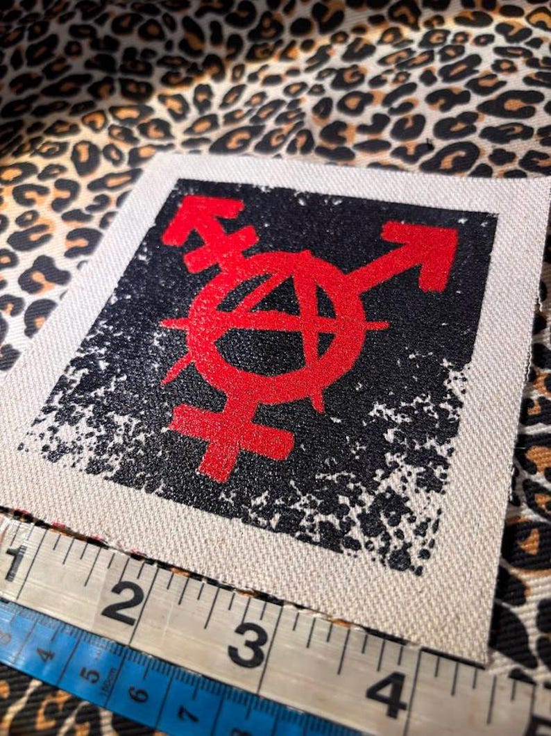 May include: A square fabric patch featuring a red anarchy symbol combined with gender symbols printed on a black distressed background. A white measuring tape is positioned along the bottom edge, showing the patch width is approximately 4 inches.