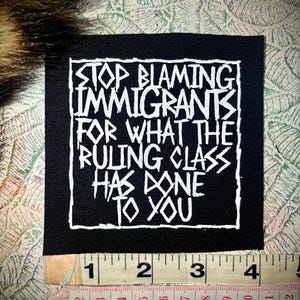 Può includere: Toppa quadrata nera con testo bianco: "STOP BLAMING IMMIGRANTS FOR WHAT THE RULING CLASS HAS DONE TO YOU". Un righello in basso mostra le misure in centimetri.