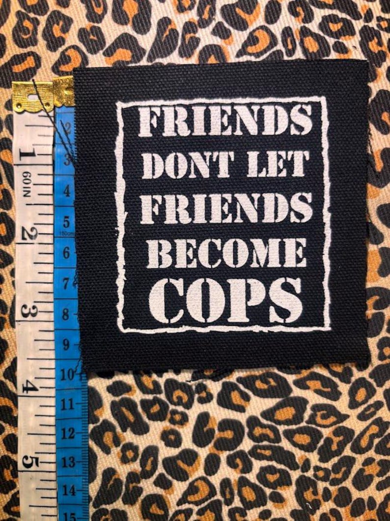 May include: A square black fabric patch with white screen-printed text that reads FRIENDS DONT LET FRIENDS BECOME COPS. The patch is positioned next to a measuring tape showing a height of approximately 4 inches or 10 centimeters.
