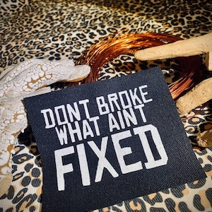 Op de afbeelding: Zwarte stoffen patch met witte tekst die "Don't broke what ain't fixed" zegt.