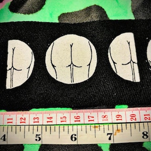 Könnte beinhalten: Ein schwarzer Stoff-Patch mit drei weißen Umrissen einer Rückansicht eines menschlichen Körpers. Der Patch wird mit einem weißen Maßband gemessen.