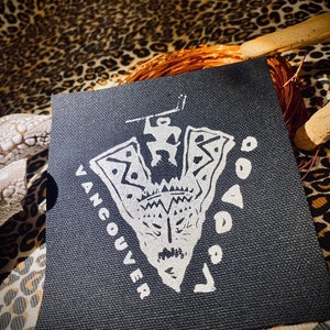 May include: Black and white embroidered patch with a stylized arrowhead design and the word "Vancouver" written around it. The arrowhead features a stylized face and a figure standing on top.