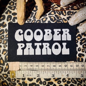May include: A black fabric patch with white lettering that reads "Goober Patrol" in a retro font style.