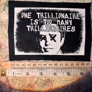 May include: A rectangular black and white fabric patch featuring a portrait with X-shaped eyes and the text ONE TRILLIONAIRE IS TOO MANY TRILLIONAIRES. A measuring tape shows the patch is approximately 6 inches wide and 4 inches tall.