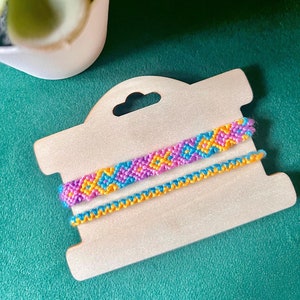 以下が含まれることがあります： 木製のディスプレイカードに2本の友情ブレスレット。上のブレスレットはピンク、ブルー、イエローで幾何学模様。下のブレスレットはイエローです。